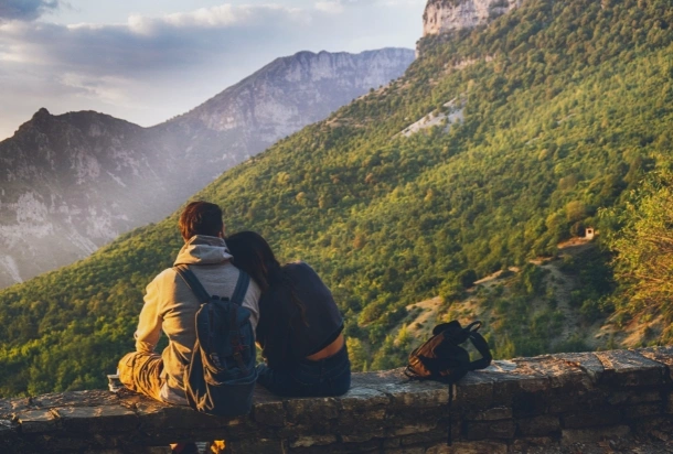 A couple sits on a stone wall, gazing at the expansive mountains in the distance under a clear blue sky.