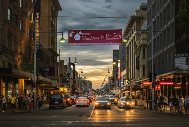 A crowded urban street in Adelaide, Australia, with vehicles and pedestrians at twilight.